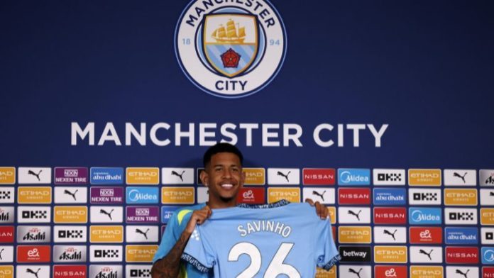 El delantero brasileño del Manchester City, Savinho, sostiene su nueva camiseta durante la presentación oficial en el estadio Etihad de Manchester.