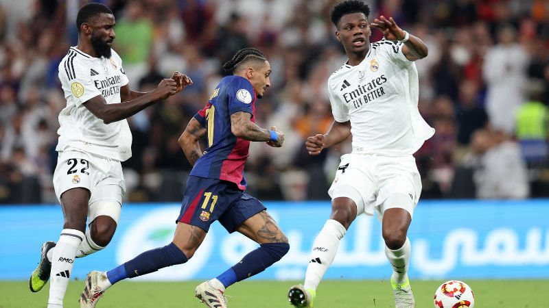 El delantero brasileño del Barcelona, ​​Raphinha, corre con el balón junto al centrocampista francés del Real Madrid, Aurelien Tchouameni, y el defensor alemán, Antonio Ruediger.