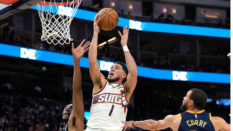 Devin Booker de los Phoenix Suns lanza sobre Kevon Looney de los Golden State Warriors.