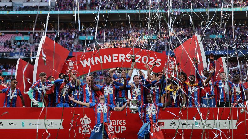 Los defensas Marc Guehi y Joel Ward, sostienen el trofeo mientras sus compañeros del Crystal Palace celebran luego de vencer al Manchester City en la final de la Copa de Inglaterra.